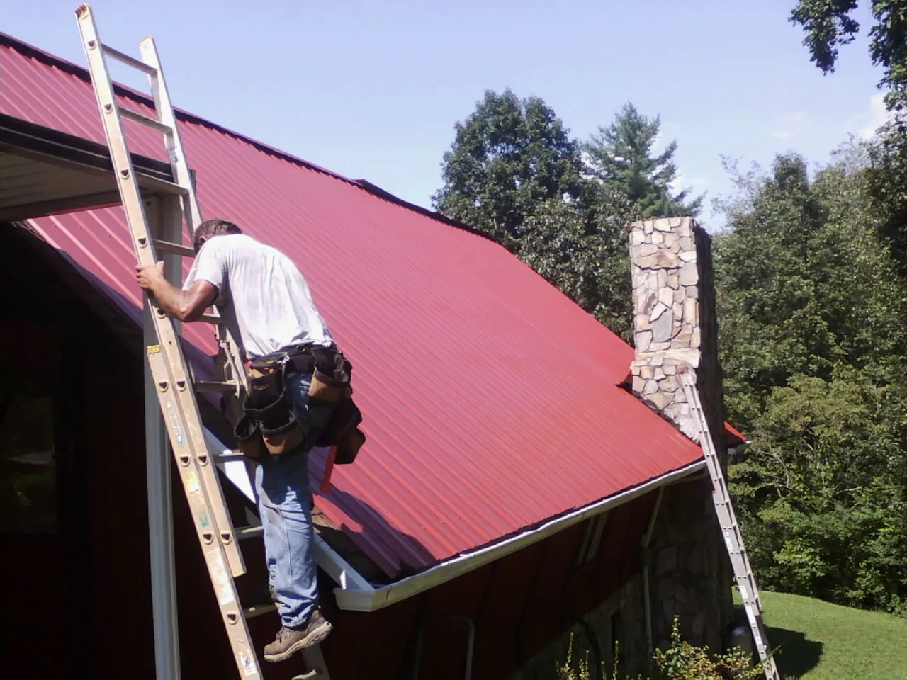 Roofer on a ladder beside a red metal roof and stone chimney — Soffit & Fascia Repair by Roofing Brothers in Gruetli Laager